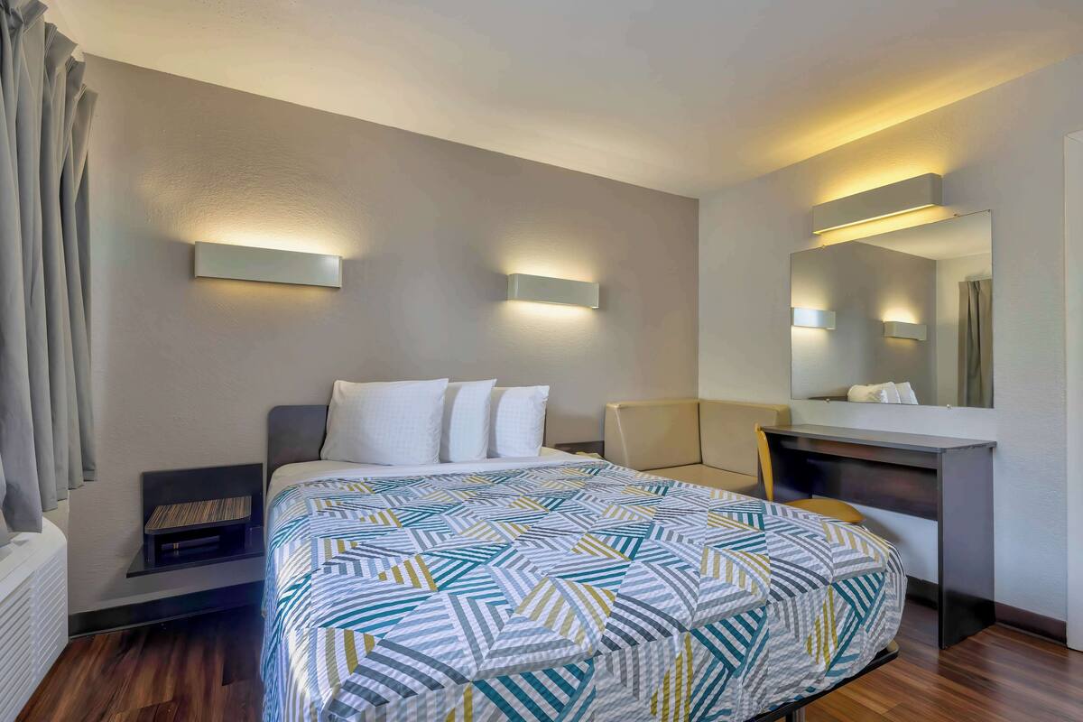 A modern hotel room with a neatly made bed, a desk with a chair, and a large mirror.