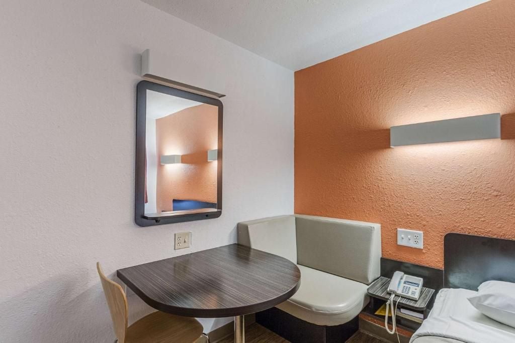 Habitación de hotel con cama, mesita de noche, silla, pequeña mesa, espejo y teléfono en la mesita de noche.