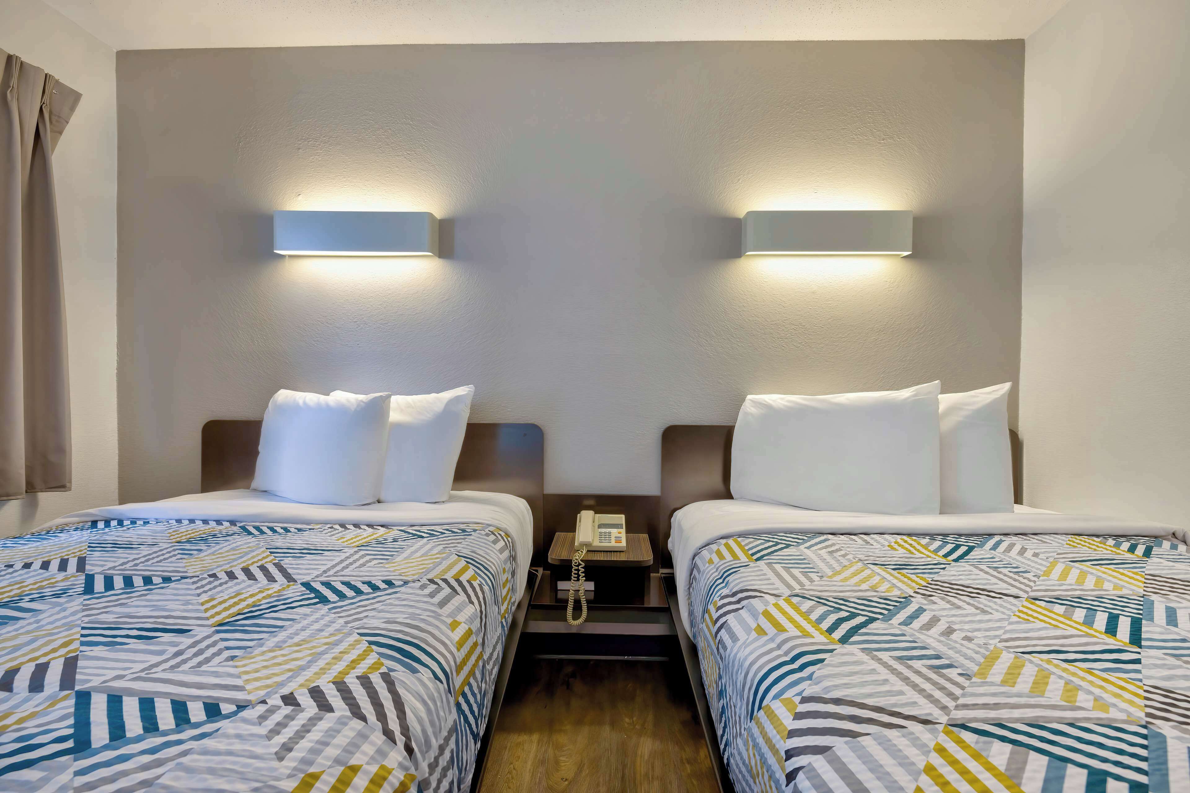 Habitación de hotel con dos camas, una mesita de noche con teléfono e iluminación en la pared.