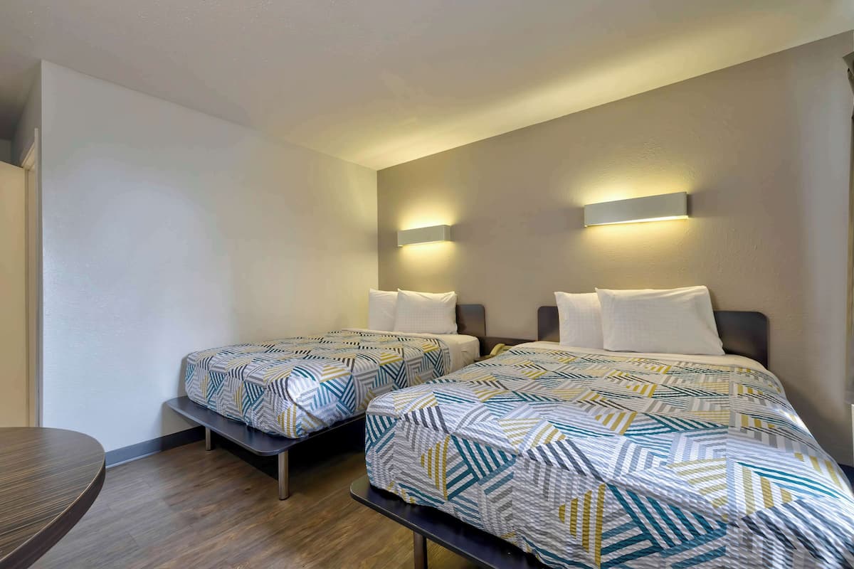 A hotel room with two beds, each with patterned bedding, a small table, and wall-mounted lighting.