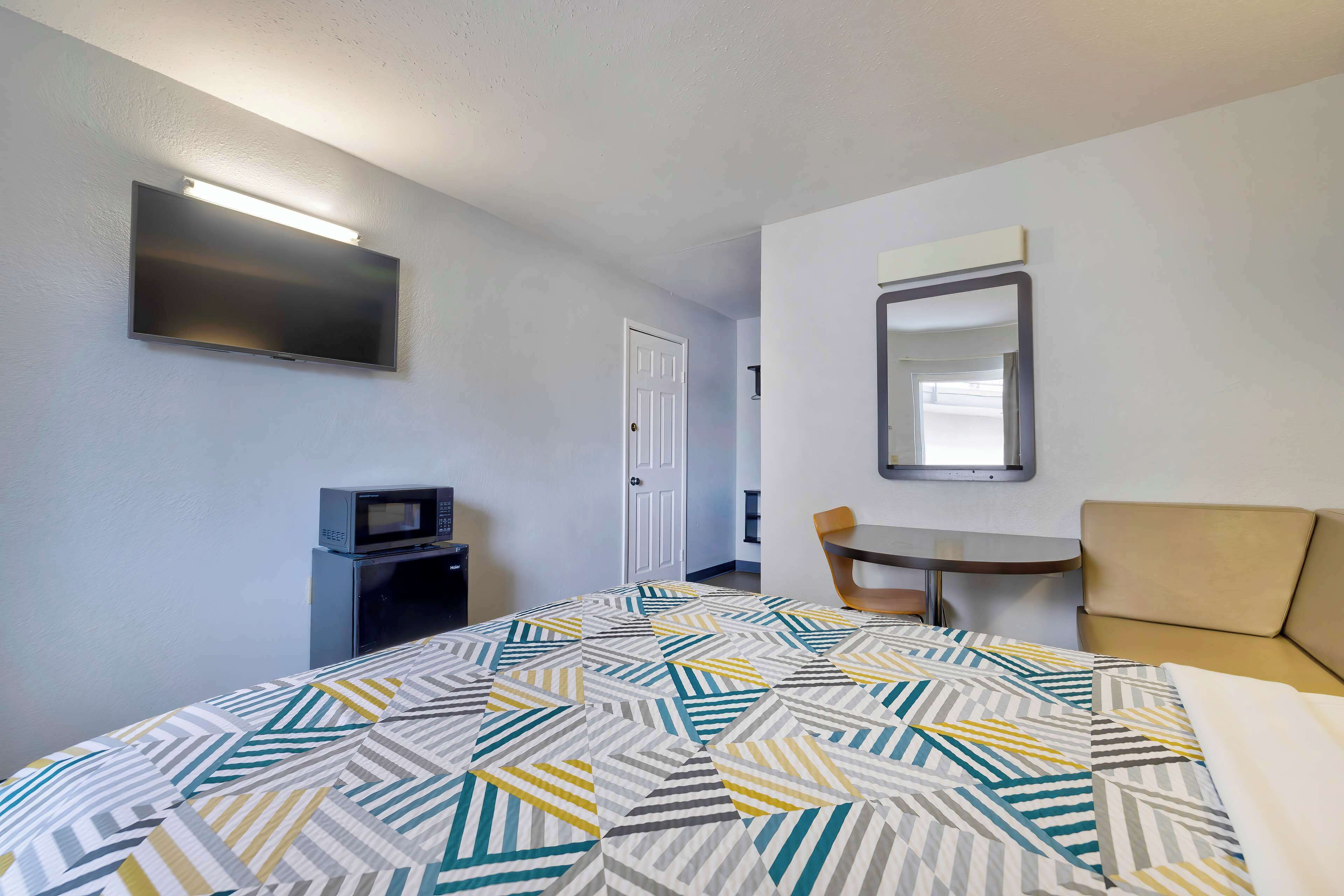 Quarto de hotel com cama, escrivaninha, cadeira, televisão e espelho.