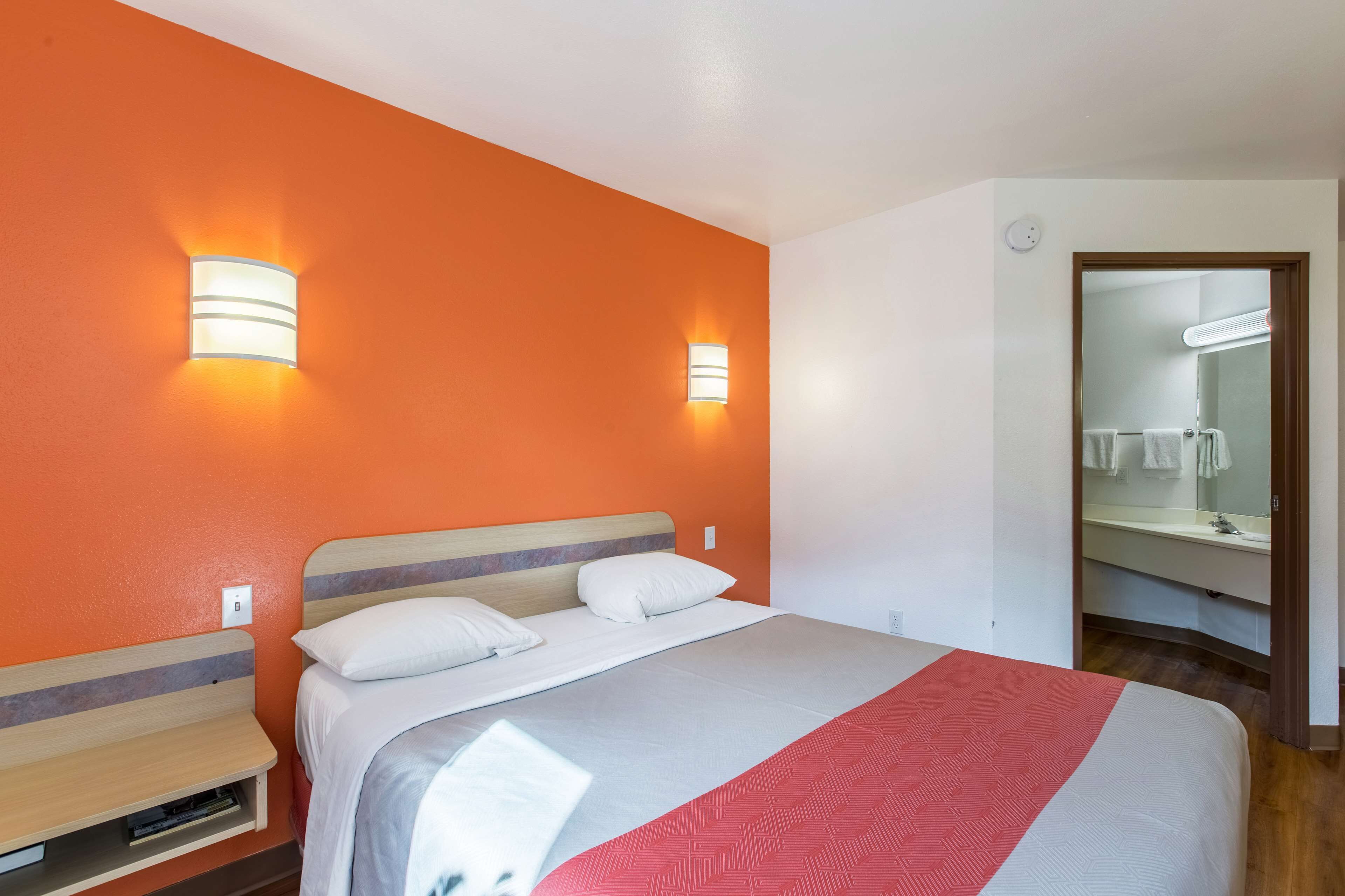 Habitación de hotel con una pared de acento naranja, una cama con ropa de cama blanca, dos mesitas de noche y un espejo sobre el lavabo.
