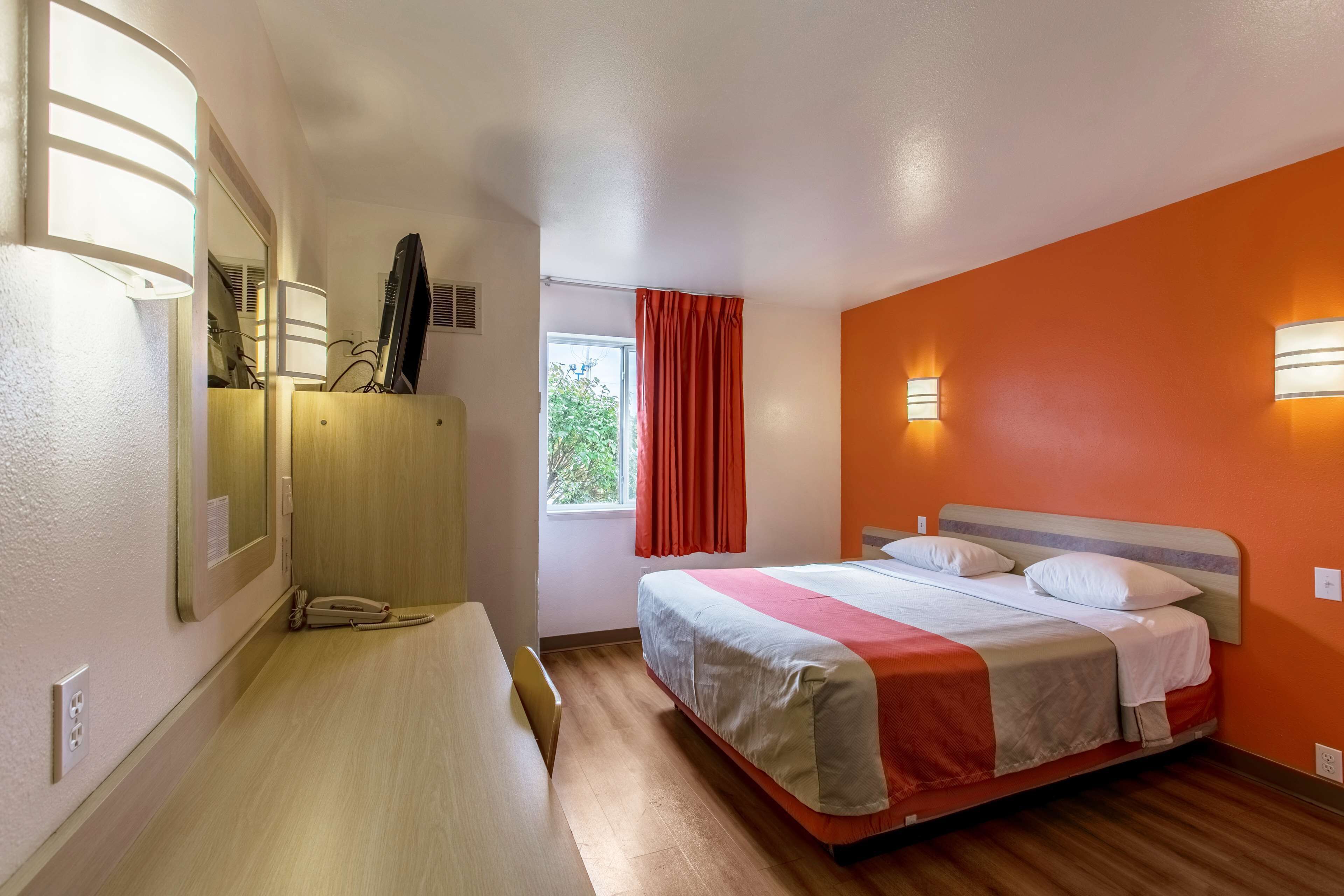 A hotel room with a bed, a desk, a chair, a window with curtains, and wall-mounted lights.