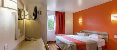 A hotel room with a bed, a desk, a chair, a window with curtains, and wall-mounted lights.