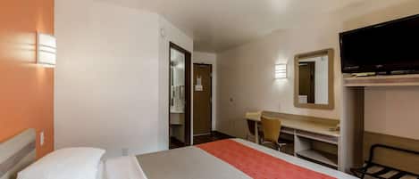 Una habitación de hotel con cama, escritorio, silla, televisor y espejo.