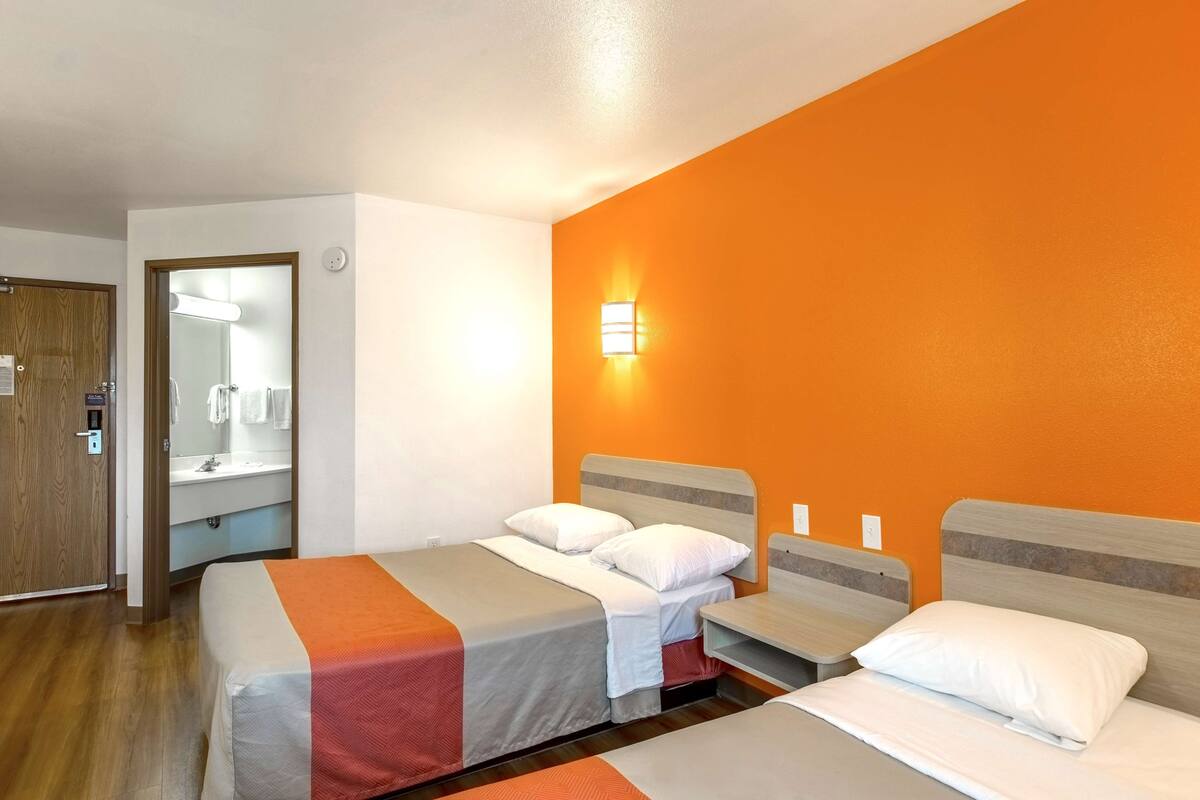 A hotel room with two beds, a bathroom with a mirror and sink, and an orange accent wall.