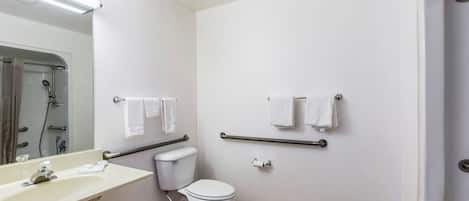 A small, clean bathroom with a white sink, toilet, and shower. There are grab bars above the toilet and a towel rack on the wall.