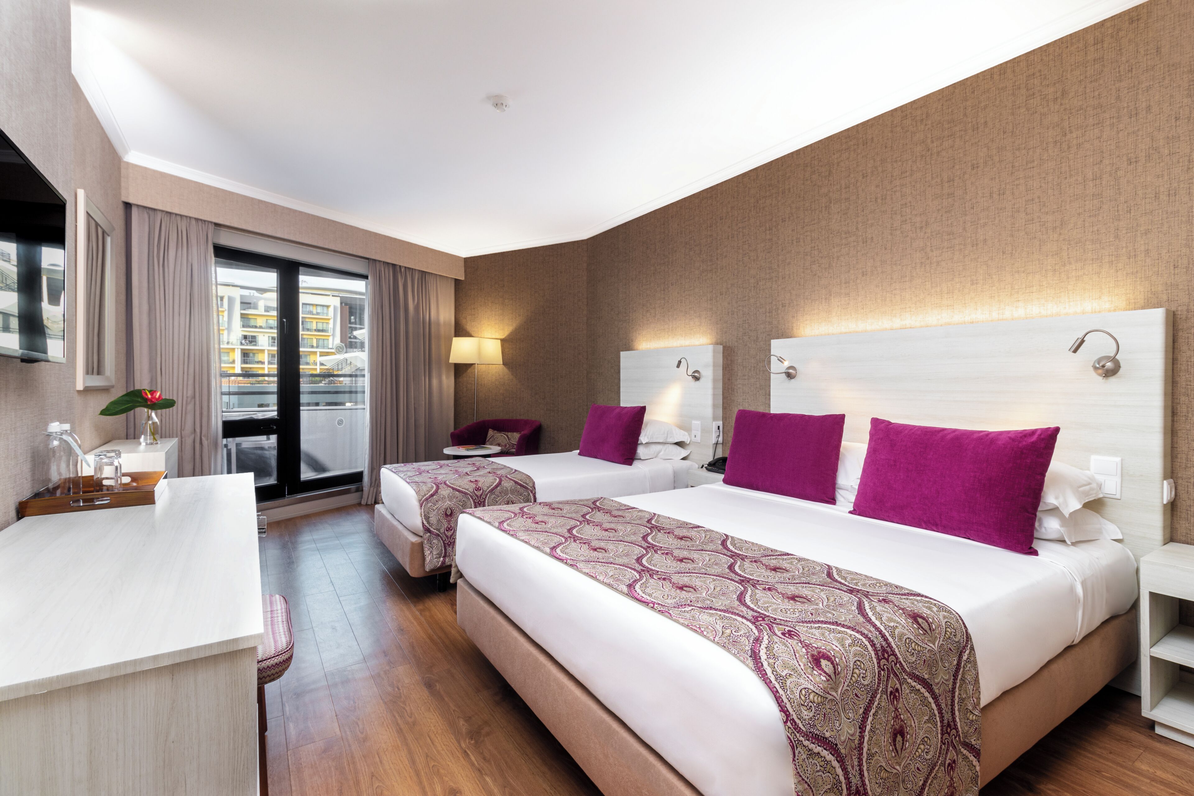 A hotel room with two beds, a desk, and a view of a building through the window.