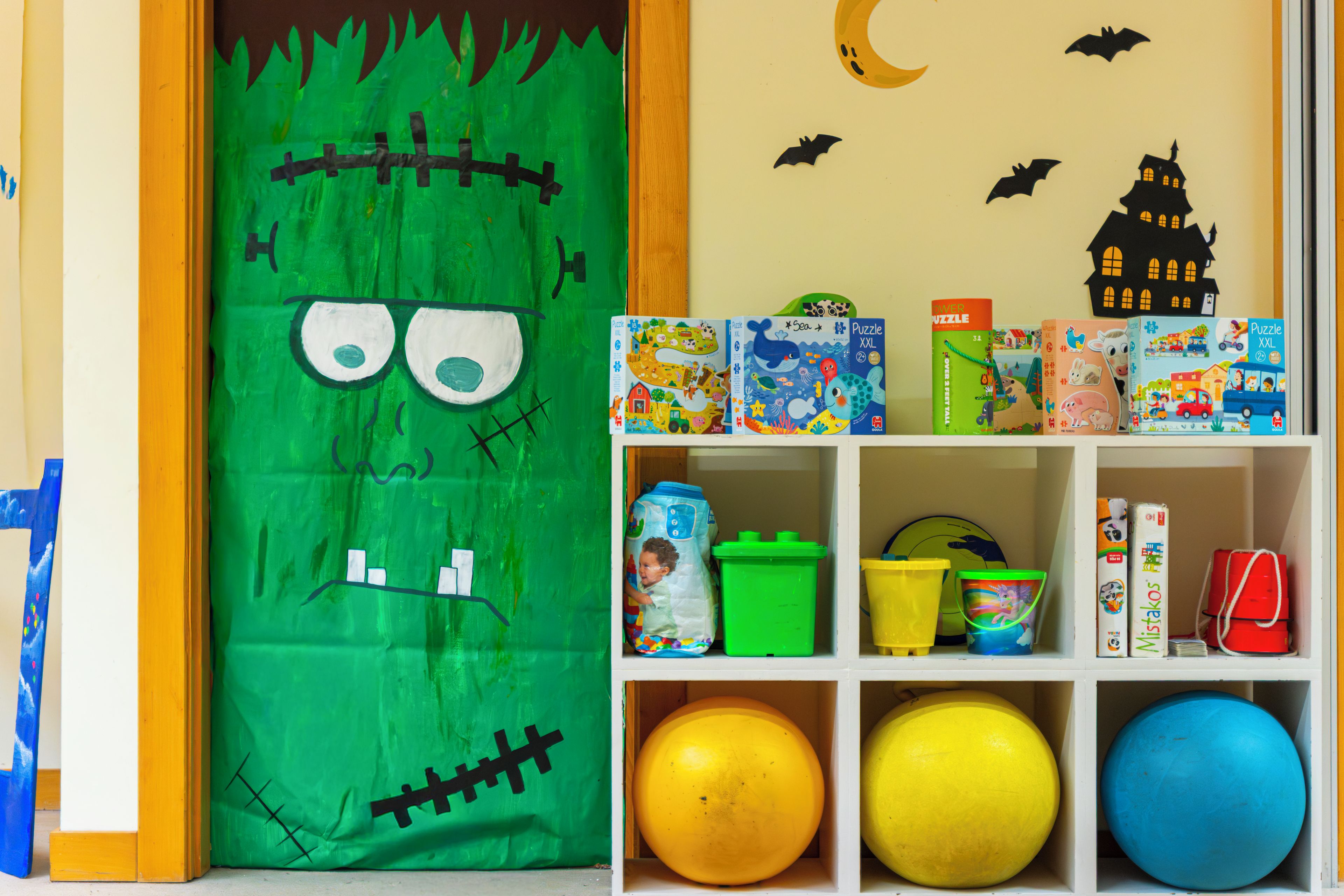 A children's room with a green door decorated with a spooky face, a shelf with various toys and books, and a wall with a moon and bats.