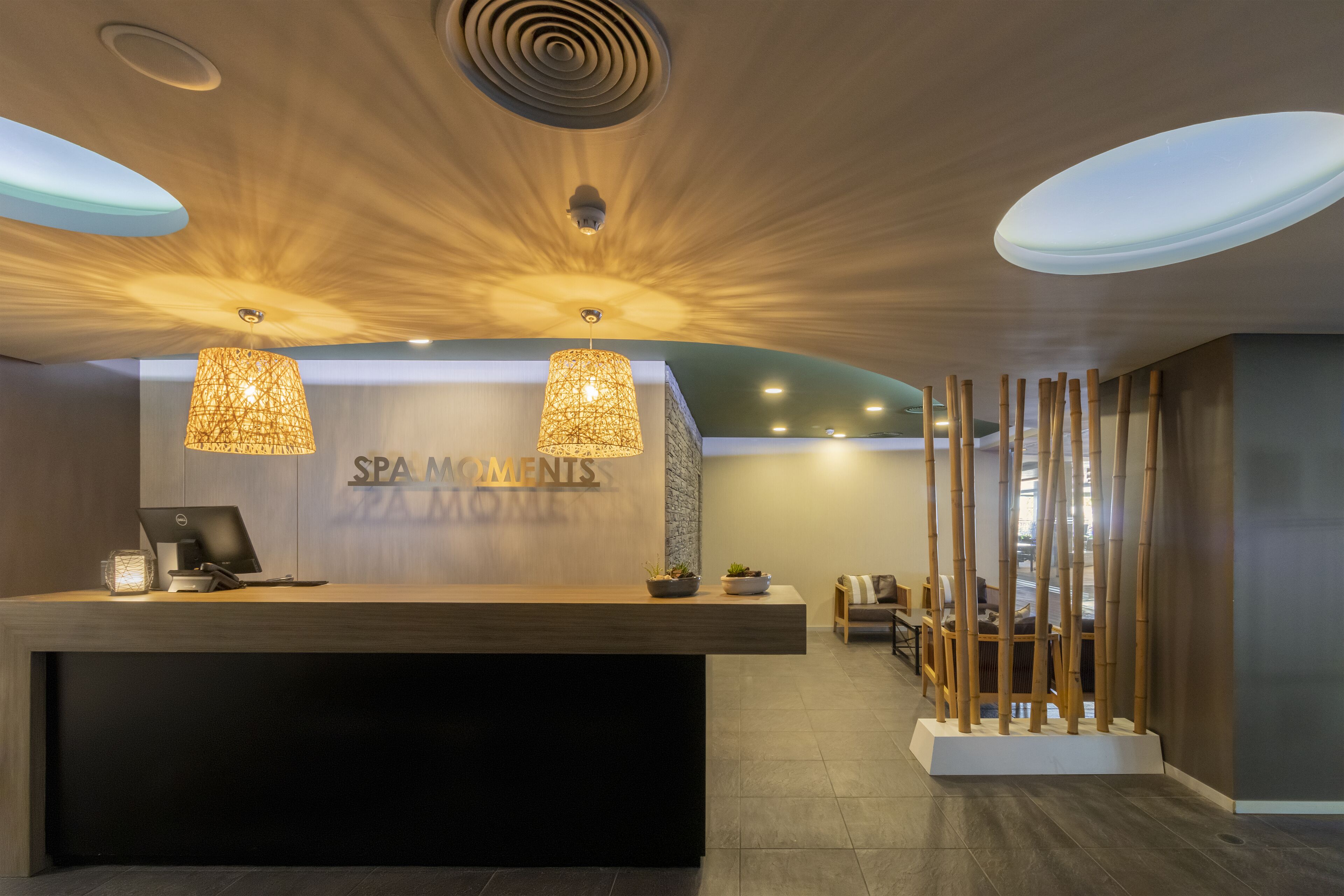 A reception area with a wooden reception desk, two pendant lights, a wall-mounted sign reading "SPA MOMENTS", and a seating area with chairs and a decorative bamboo structure.