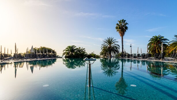 Une piscine à débordement spacieuse et d’une grande beauté, bordée de chaises longues et d’ombrelles, et entourée de palmiers sous un ciel dégagé.