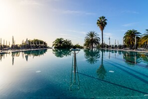 A clear, expansive infinity pool with lounge chairs and umbrellas, surrounded by palm trees and a clear sky. -  Enotel Lido – All Inclusive (Funchal)
