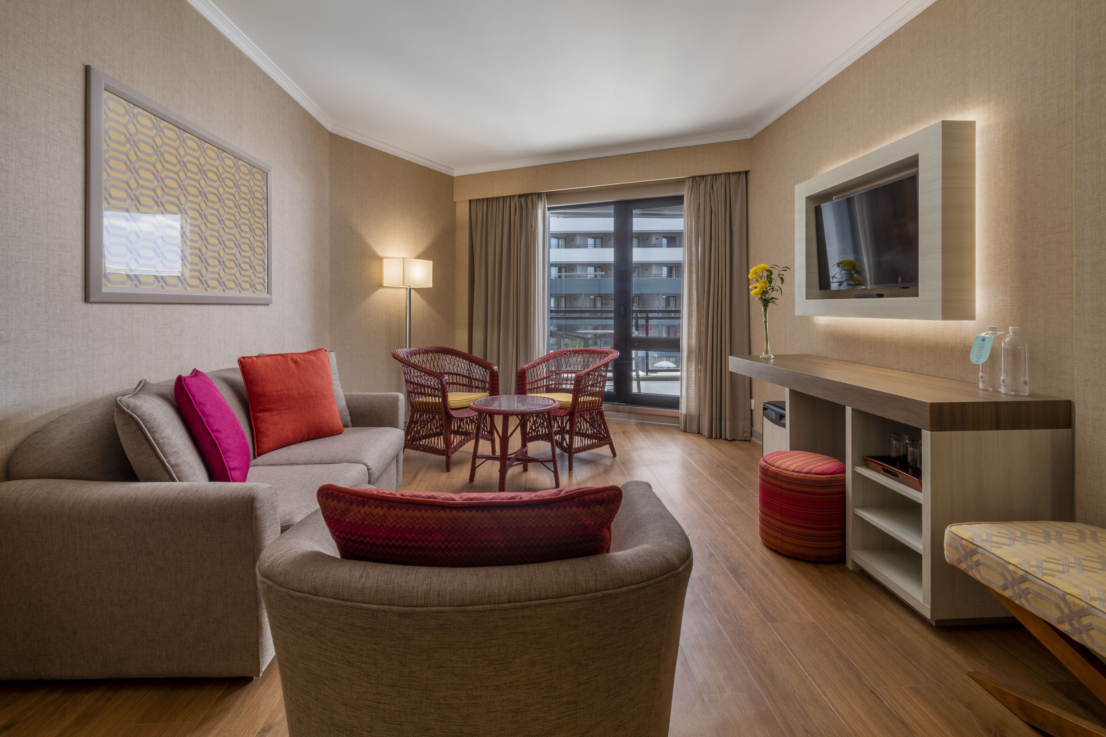 A modern hotel room with a sofa, armchair, dining table, and chairs. There is a TV mounted on the wall, a lamp, and a window with curtains.