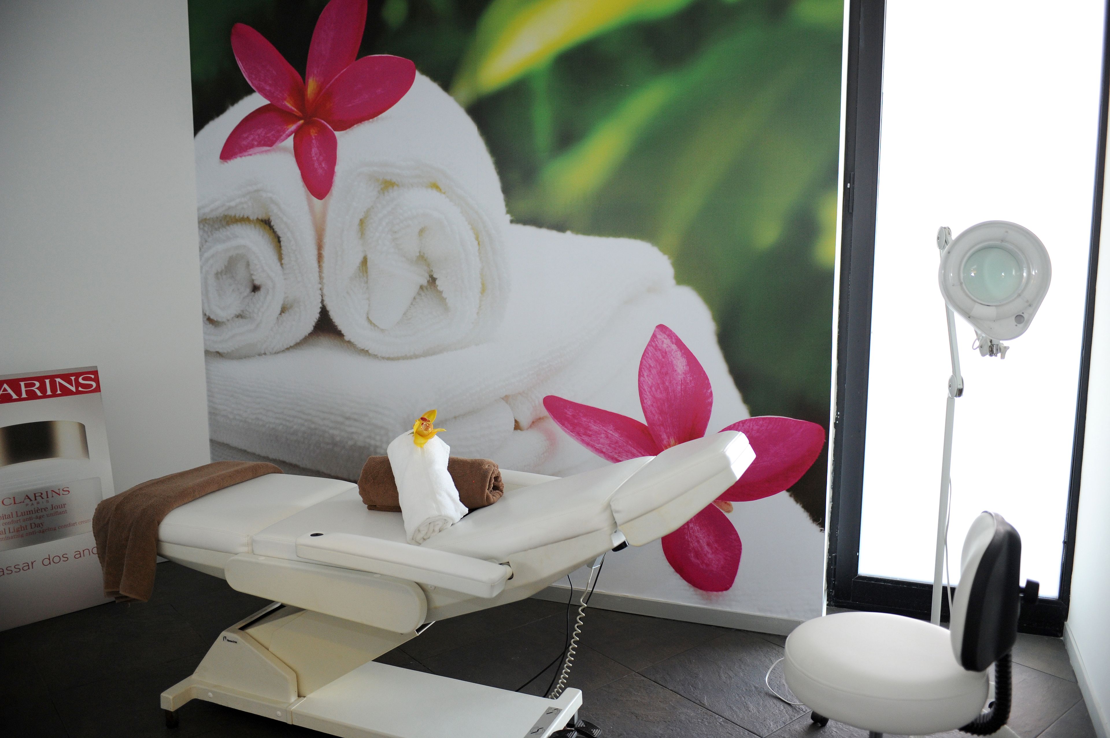 A medical examination room with a white examination table, a white chair, a large poster of towels and flowers, and a medical light.