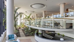 Un hall d’hôtel moderne avec un pont de verre, des palmiers et une fontaine centrale.