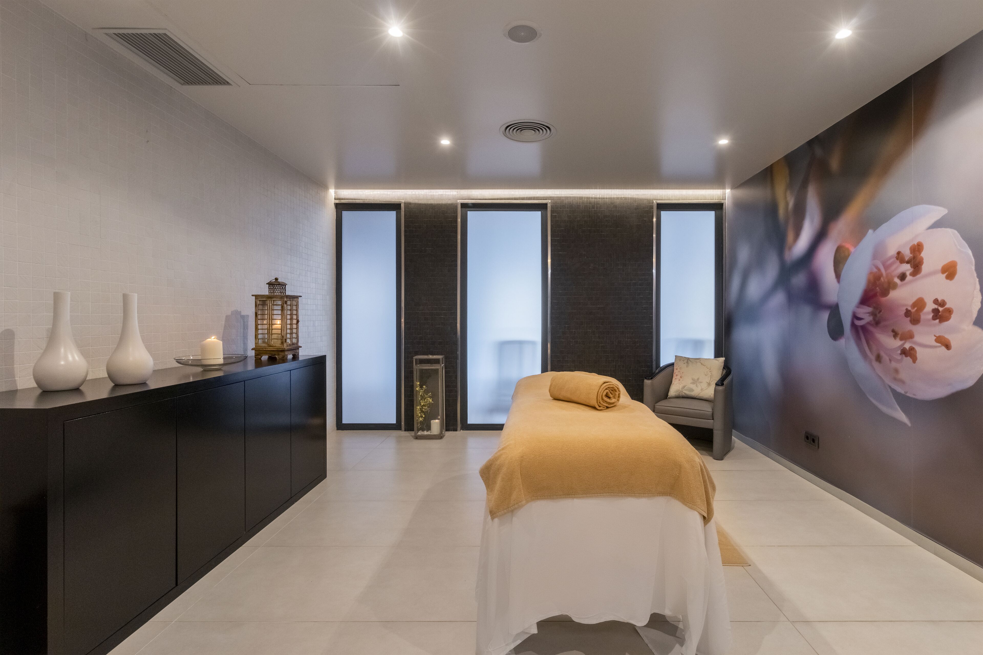 A modern spa room with a bed, a dark wooden cabinet, and a large floral mural on the wall.