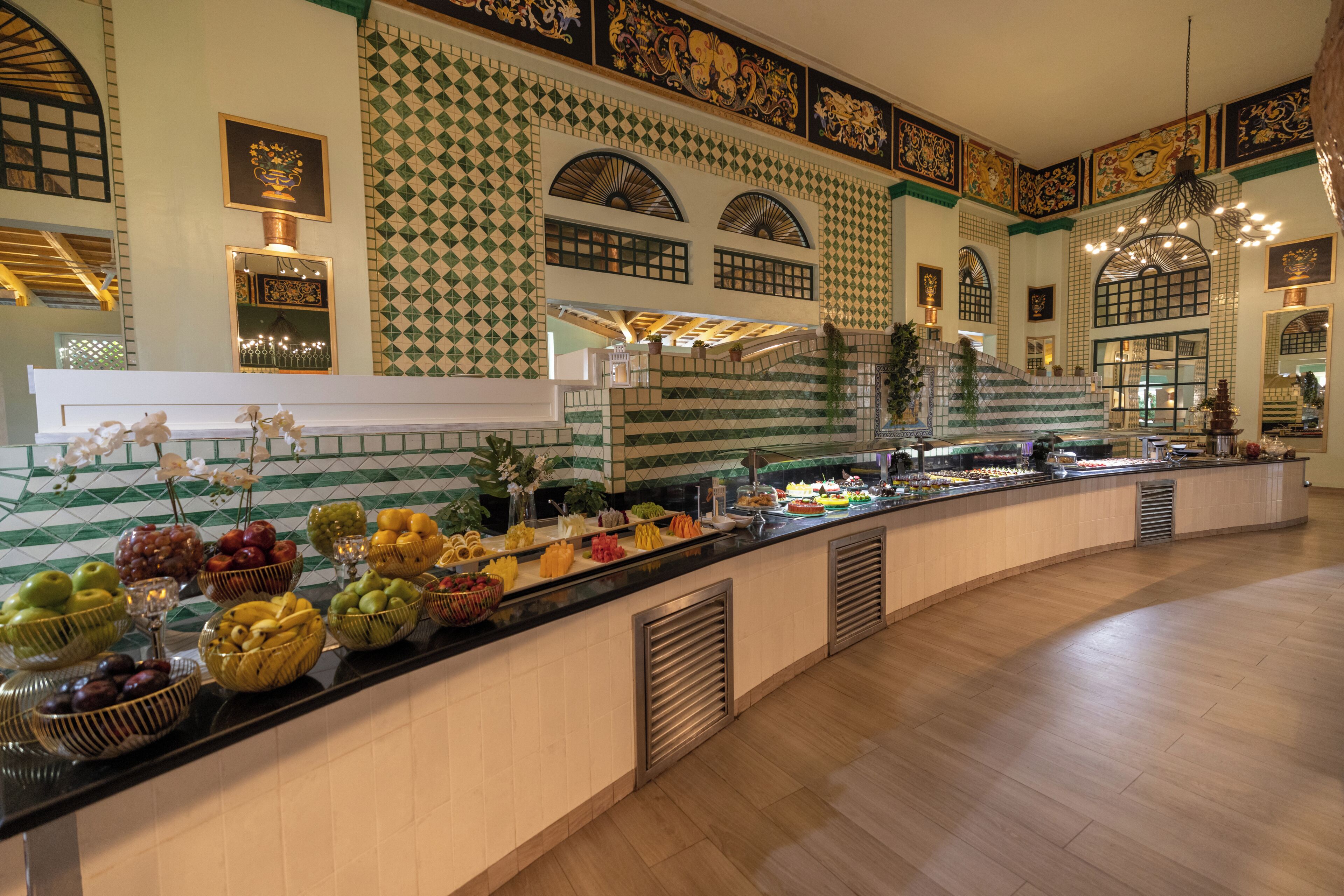 A buffet with various fruits and vegetables, a white tiled wall with green patterns, and a staircase leading to an upper level.