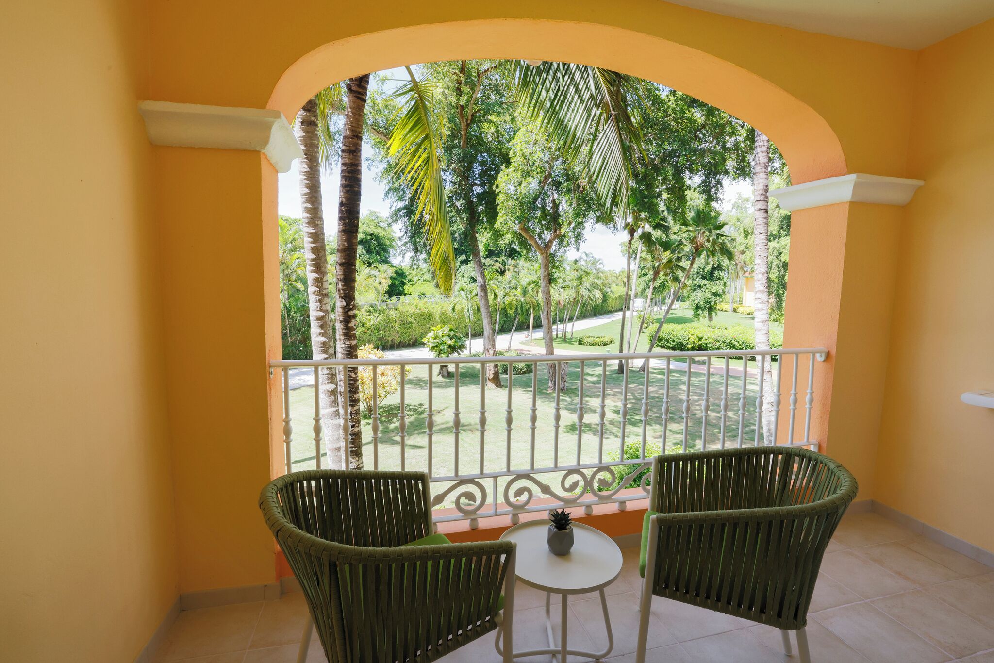 Un balcon agrémenté de deux fauteuils en osier et d’une petite table, donnant sur un jardin bien entretenu, avec divers arbres et une pelouse soigneusement taillée.