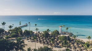 A beach with palm trees, a pier, and a building with a striped roof. - Iberostar Selection Hacienda Dominicus - All Inclusive (San Rafael del Yuma)