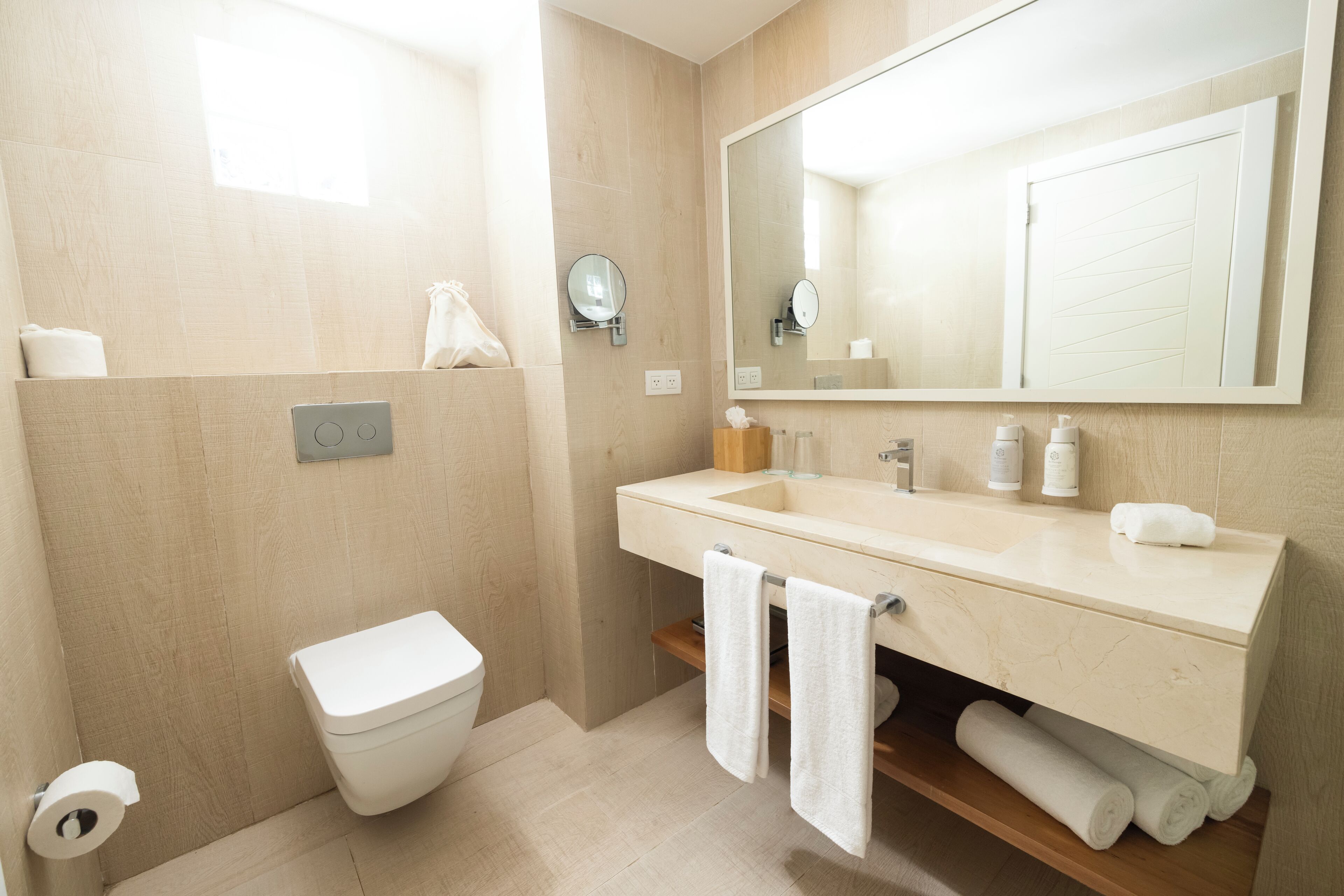 A modern bathroom with a beige sink, mirror, and toilet. There are towels hanging on the wall and a toilet paper roll on the floor.