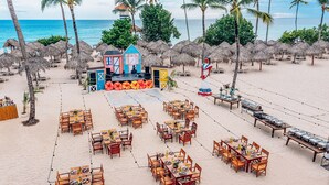 An outdoor dining area with tables and chairs arranged on a sandy beach, surrounded by palm trees and thatched umbrellas. - Iberostar Selection Hacienda Dominicus - All Inclusive (San Rafael del Yuma)