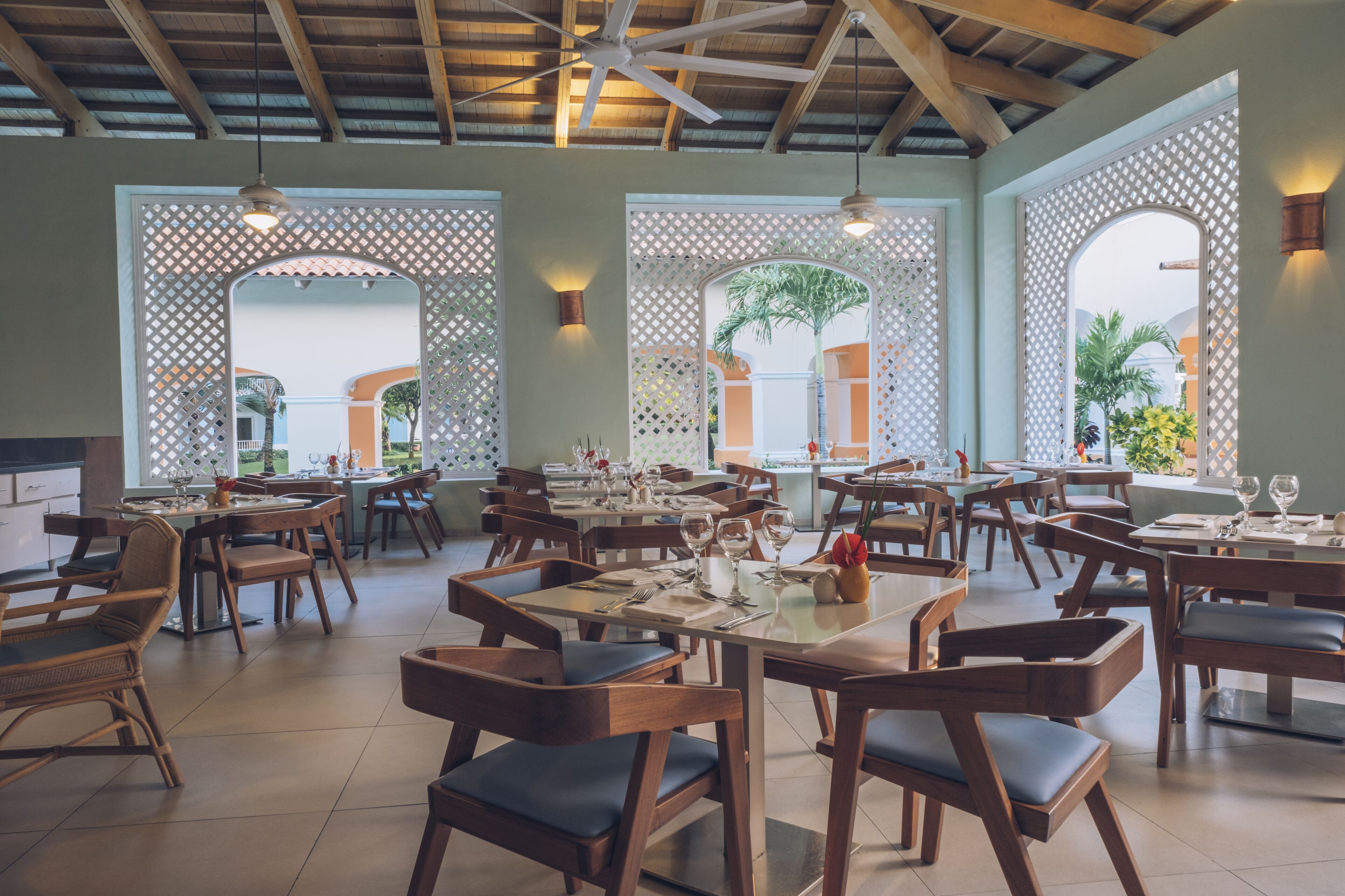 A spacious restaurant with multiple tables and chairs, wooden beams on the ceiling, and large windows with views of the outdoors.