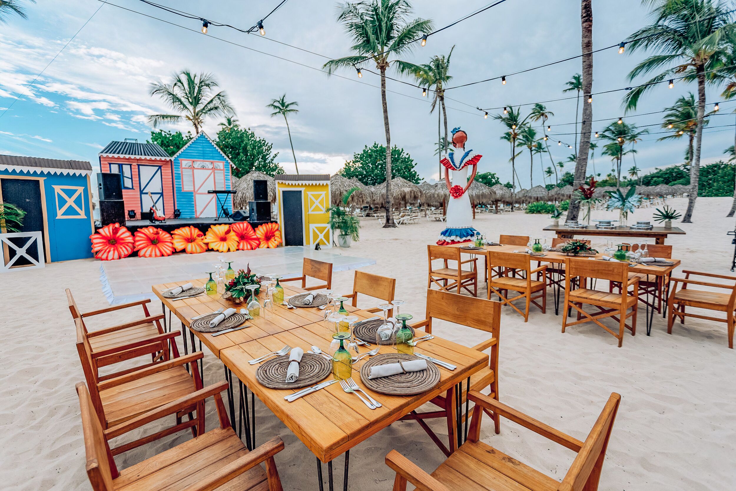 An outdoor dining area with wooden tables and chairs, set against a beach backdrop with palm trees and a stage with colorful decorations.