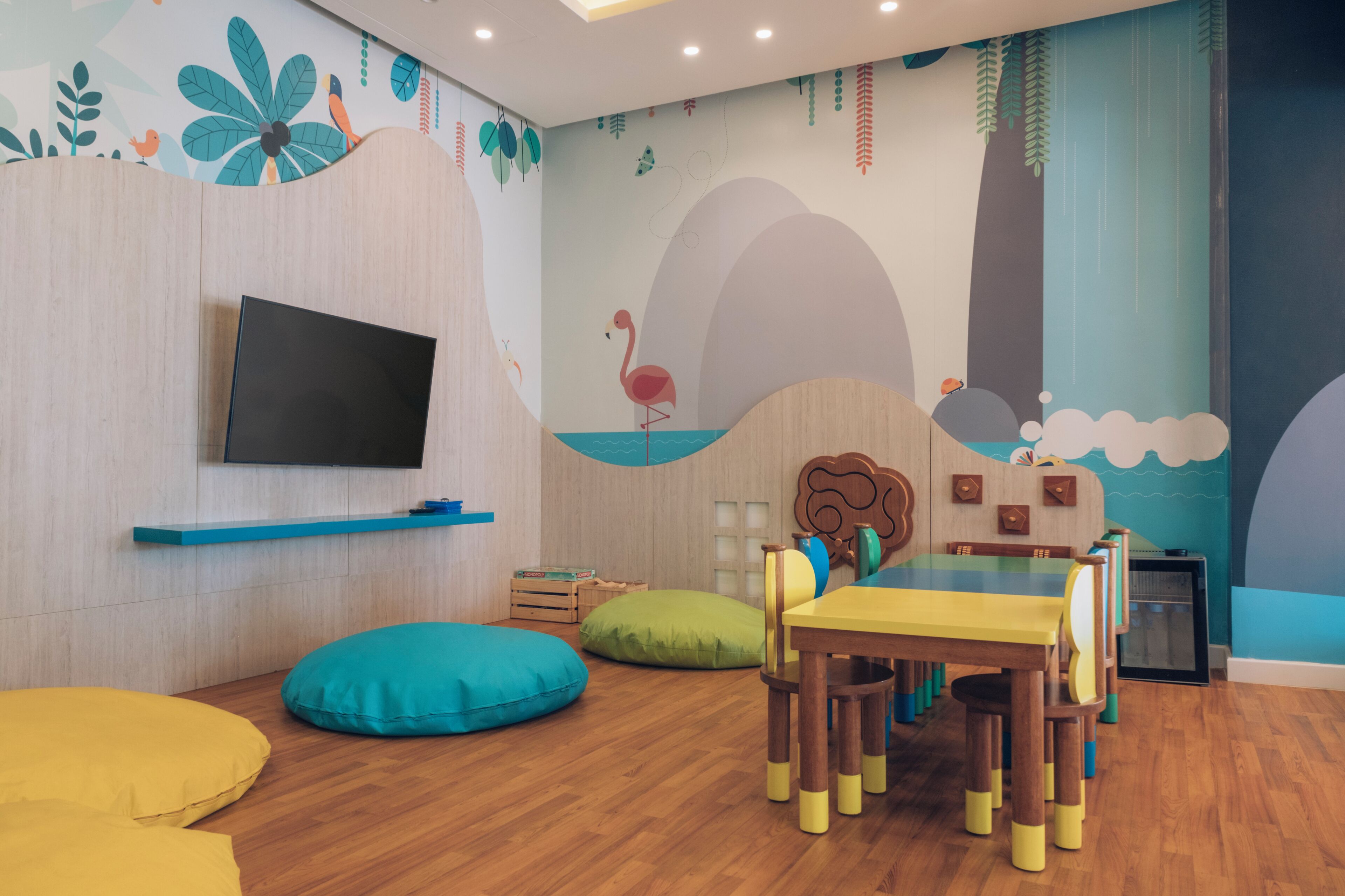Una sala de juegos para niños, con suelo de madera, televisor montado en la pared, una mesa con sillas y cojines de colores.