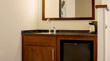A hotel bathroom with a wooden vanity, a sink, a mirror, and a refrigerator.