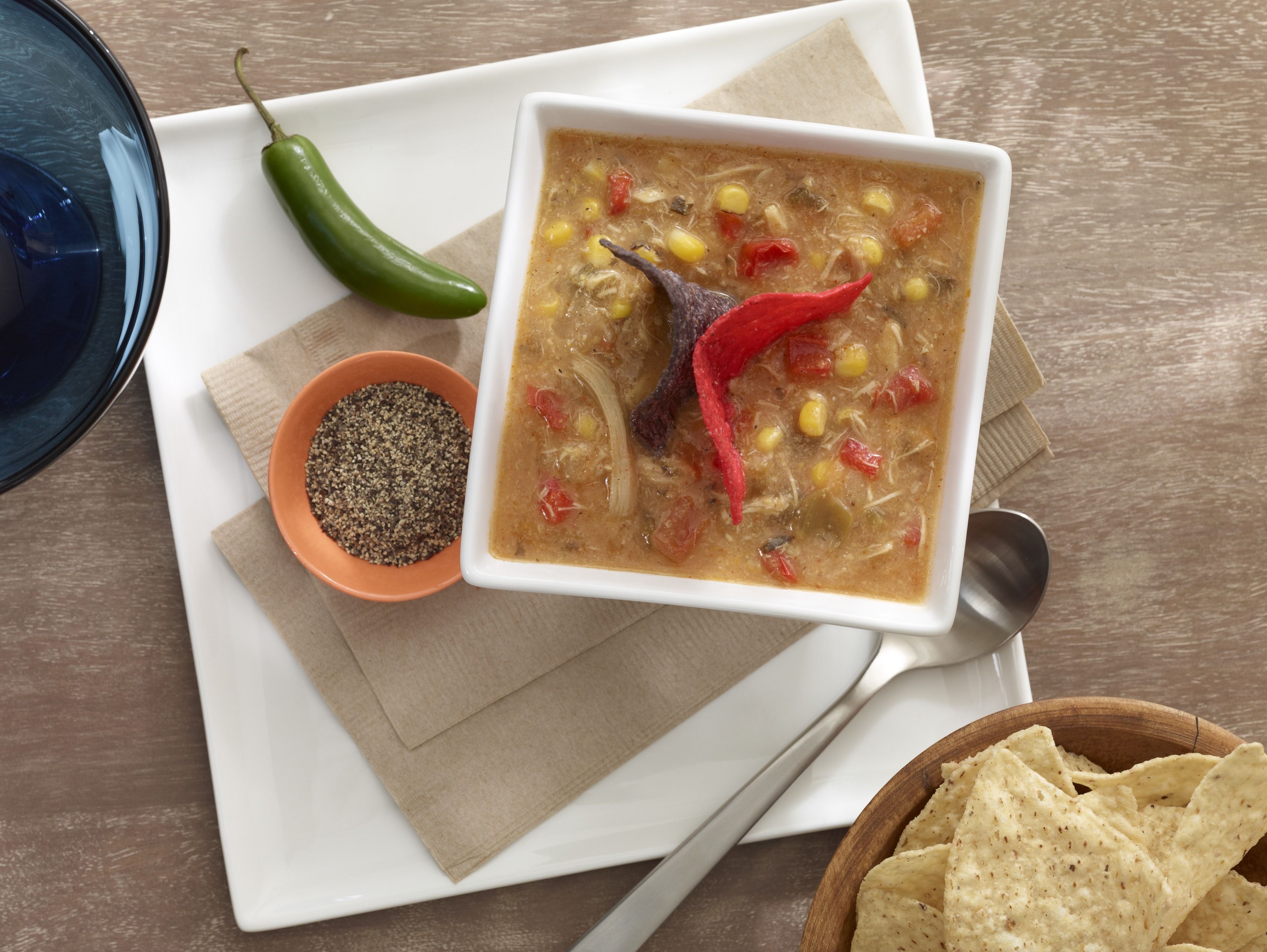 Un tazón de sopa con un chile rojo, un recipiente pequeño de especias y totopos sobre una mesa de madera.