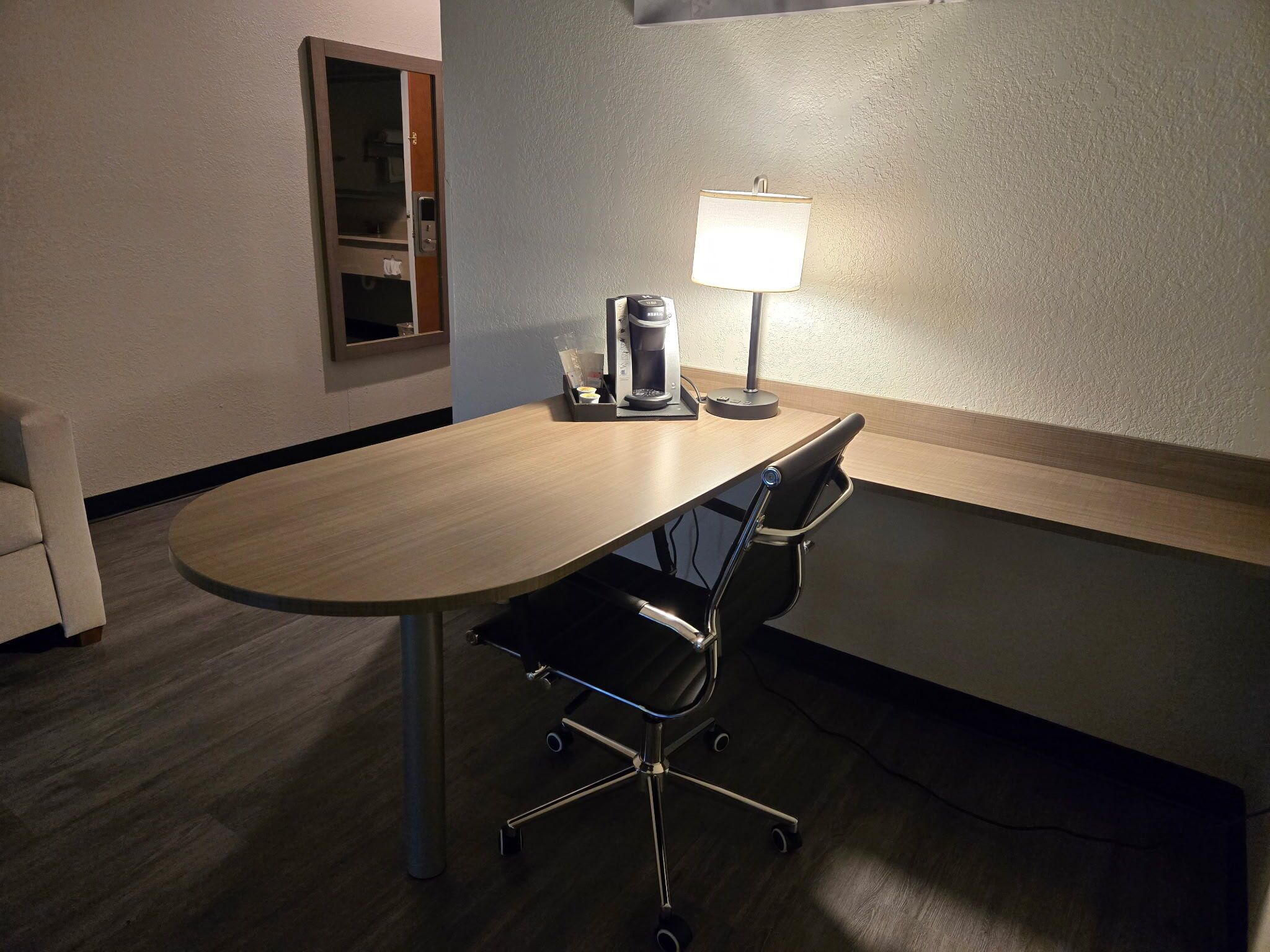 A hotel room with a round table, a coffee maker, a lamp, a chair, and a window.