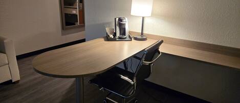 A hotel room with a round table, a coffee maker, a lamp, a chair, and a window.