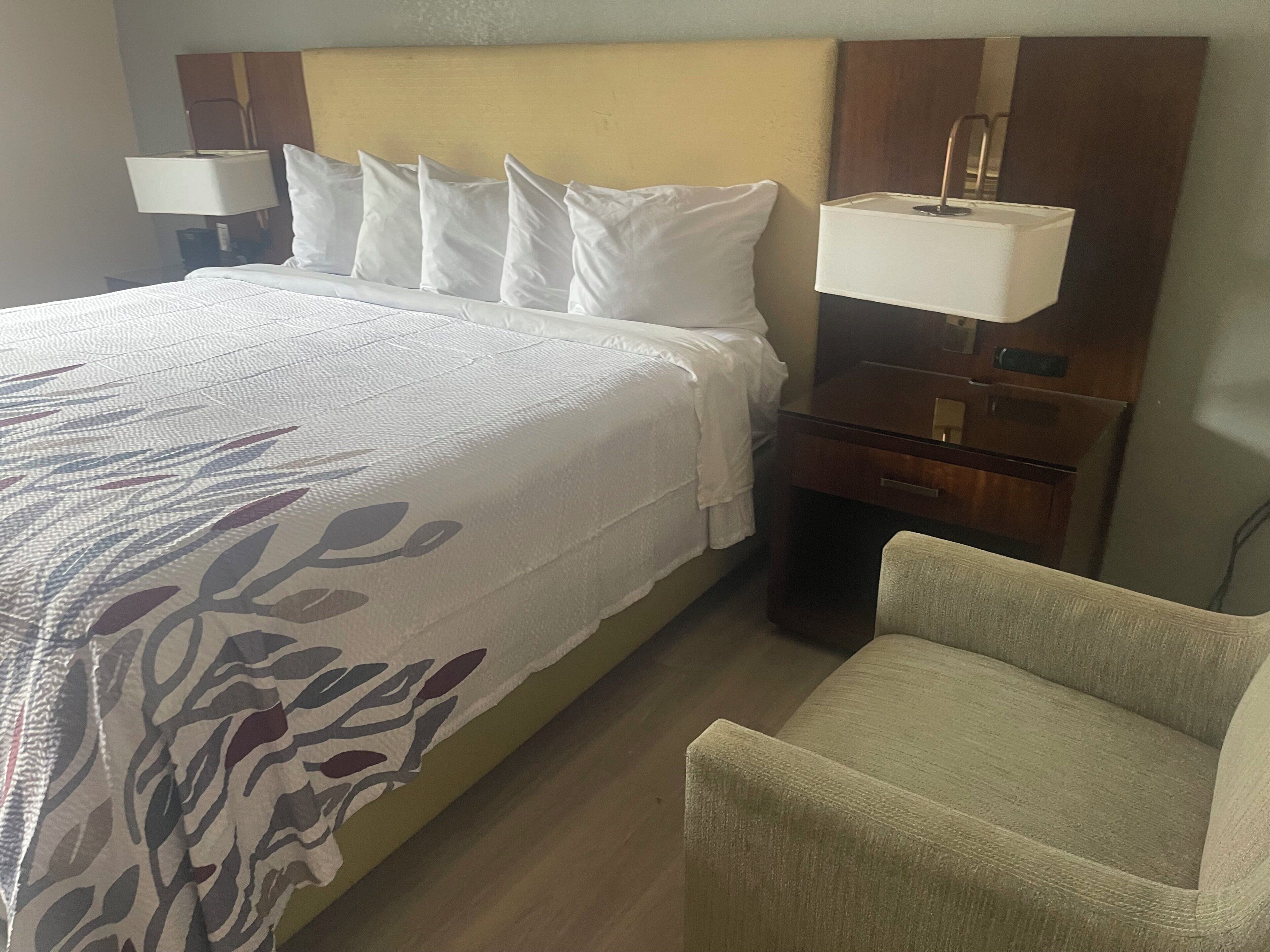 Una camera d'albergo con un letto rifatto, un comodino con una lampada, una sedia e una piccola scrivania anch'essa illuminata.