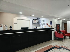 Zona de recepción de un hotel con un mostrador negro, un televisor montado, certificados enmarcados y sillas rojas.
