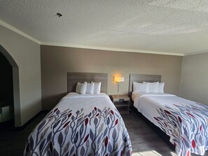 Ein Hotelzimmer mit zwei Betten, jeweils mit einem gemusterten Überwurf, einem Nachttisch, einer an der Wand befestigten Lampe und einer Deckenleuchte.