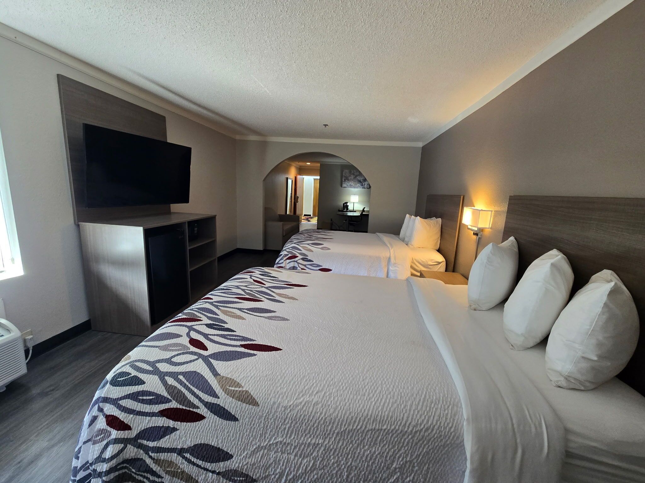 A hotel room with a large bed, a TV mounted on the wall, a desk with a lamp, and an arched doorway leading to another room.
