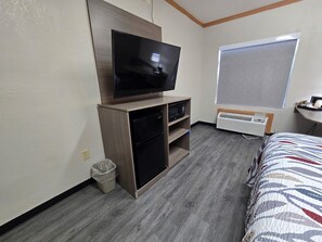 A hotel room with a flat-screen TV on a wooden stand, a small refrigerator, a waste bin, a window with a view, and a bed with a patterned bedspread. - Red Roof Inn & Suites Hinesville (Hinesville)
