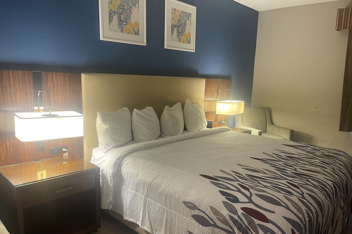Habitación de hotel con una cama grande, dos mesitas de noche con lámparas, una mesita auxiliar de madera y dos obras de arte enmarcadas en la pared.