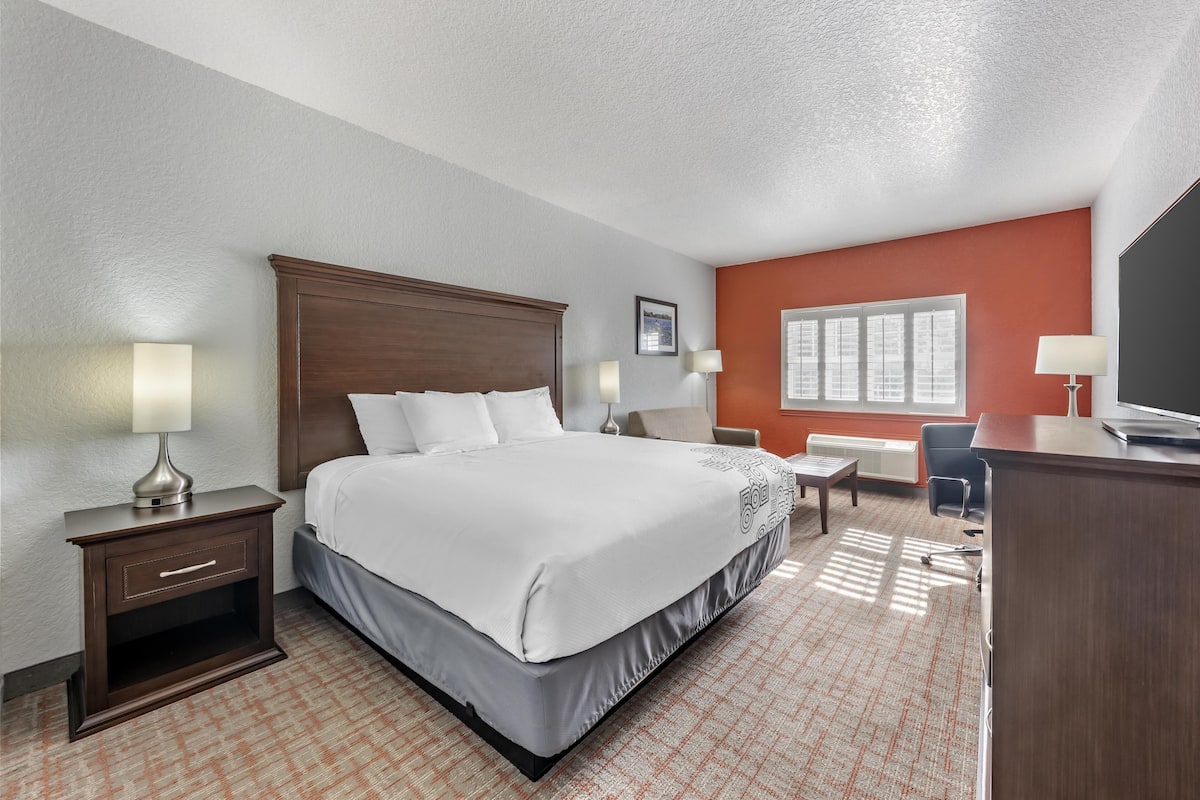 A hotel room with a large bed, a desk, a chair, a nightstand, and a window with blinds.