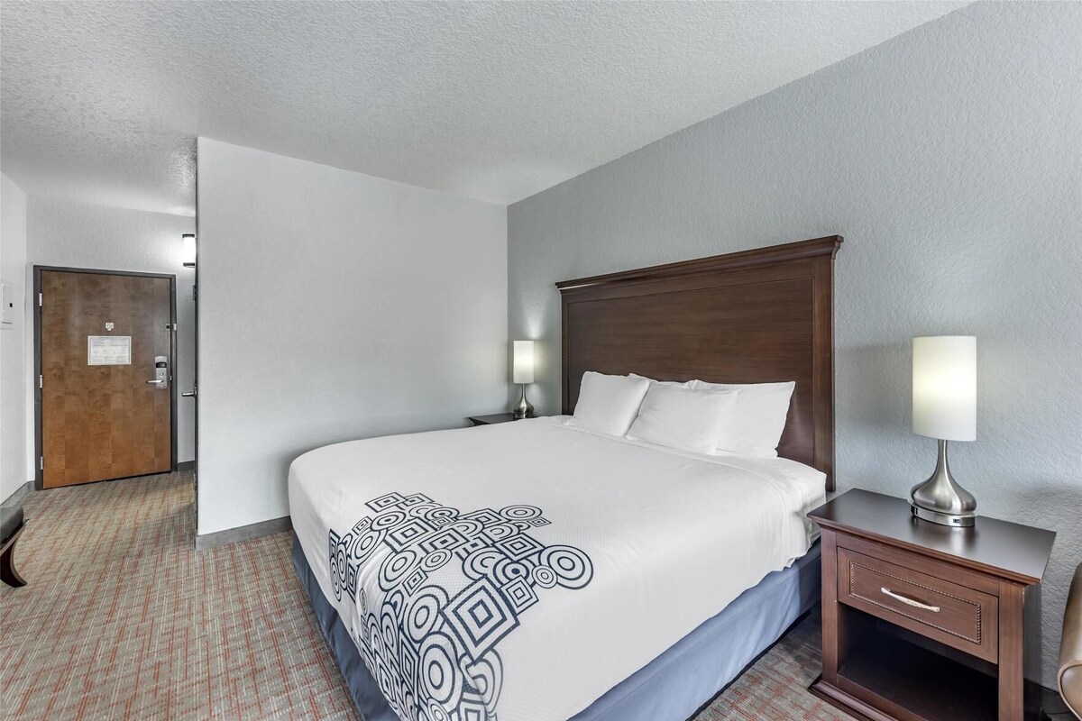 A hotel room with a large bed, a wooden headboard, a nightstand with a lamp, a door, and a carpeted floor.