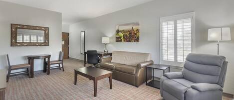A living room with a sofa, armchair, coffee table, and chairs. There is a painting on the wall and a window with blinds.