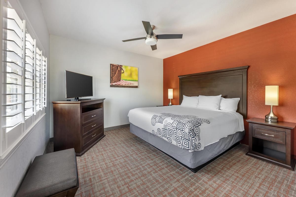 A hotel room with a bed, a TV, a desk, a chair, a nightstand, a lamp, a ceiling fan, and a window with blinds.