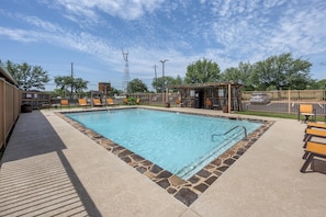 Una piscina rectangular con bordes de piedra, rodeada de sillas naranjas y una zona de estar cubierta.
