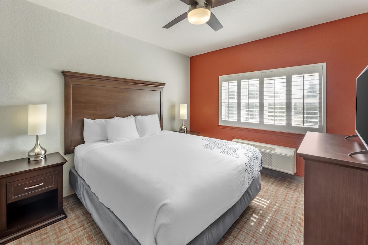 A hotel room with a large bed, a nightstand with a lamp, a desk with a television, and a window with blinds.