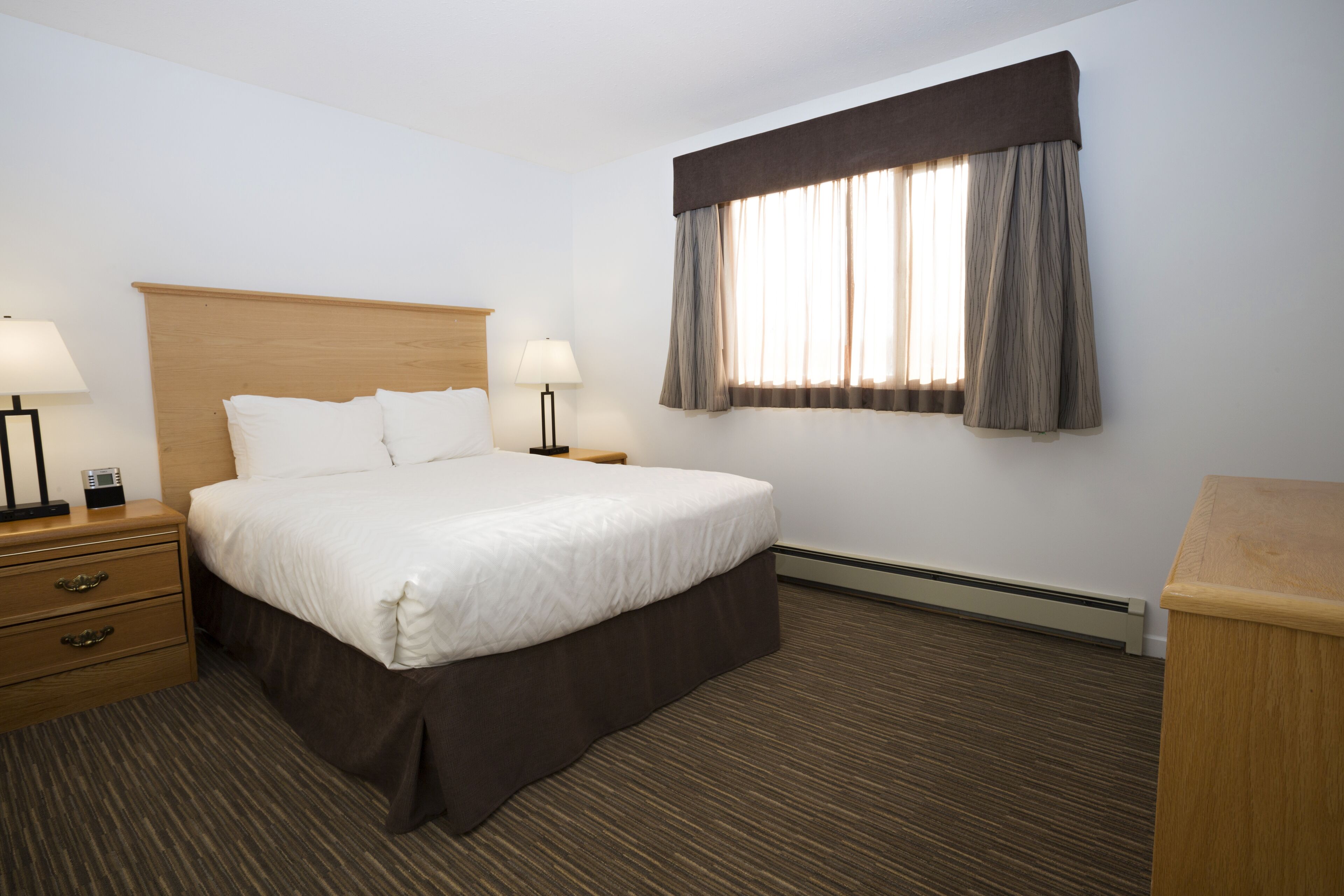 A hotel room with a large bed, two bedside tables with lamps, a dresser, and a window with curtains.