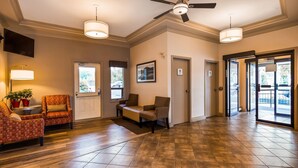A hotel lobby with a wooden floor, two seating areas with chairs, a ceiling fan, and a large window. - Tower Inn and Suites (Quesnel)