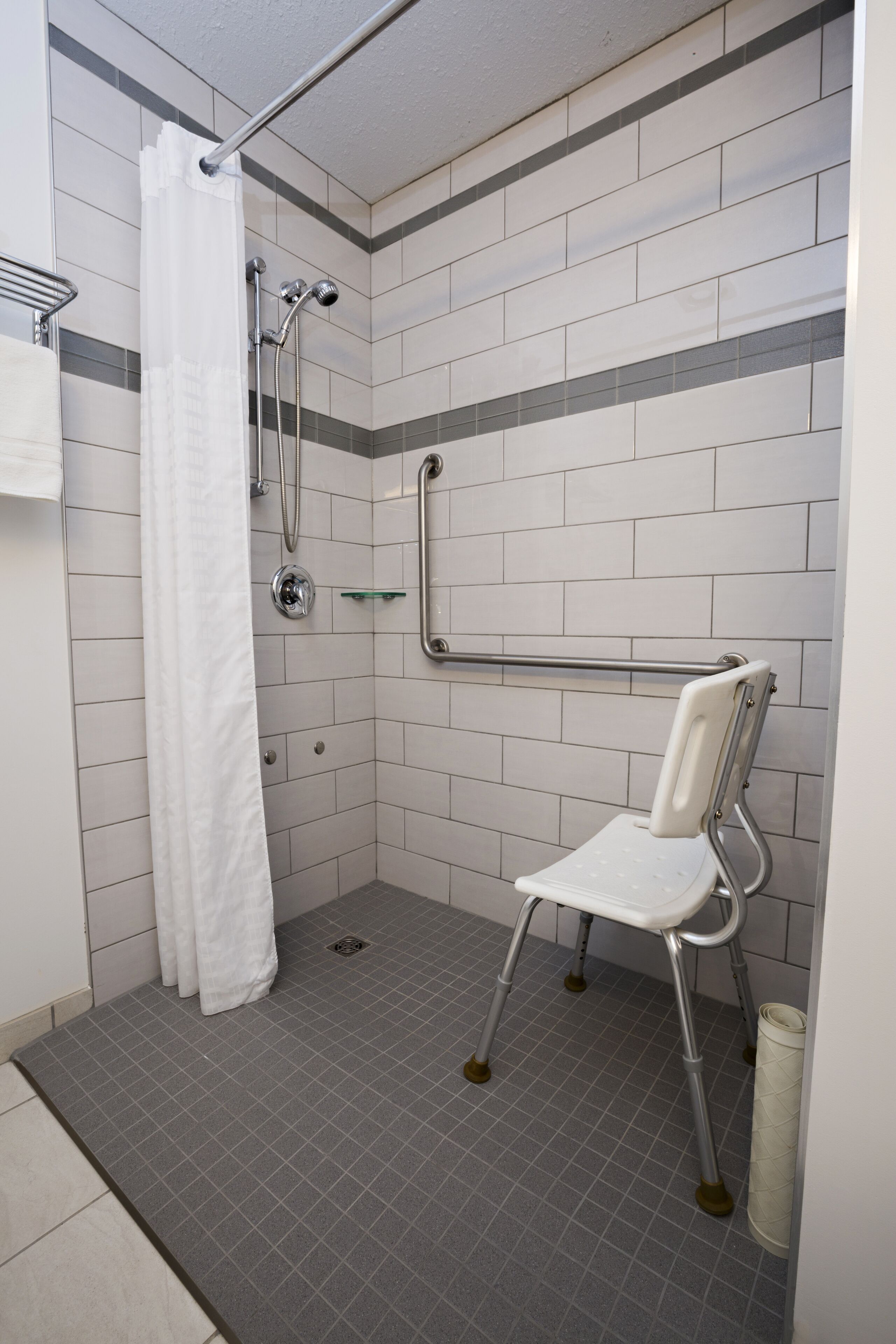 A shower area with a white shower curtain, a grab bar, and a white chair.