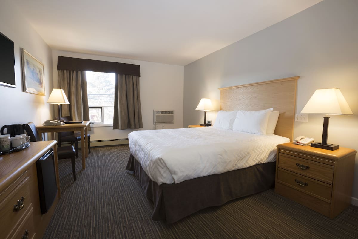 Una habitación de hotel con una cama grande, dos mesitas de noche con lámparas, un escritorio con una silla, un aparador y una ventana con cortinas.