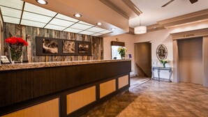 A hotel reception area with a wooden reception desk, framed artwork, and a staircase leading to an elevator. - Tower Inn and Suites (Quesnel)