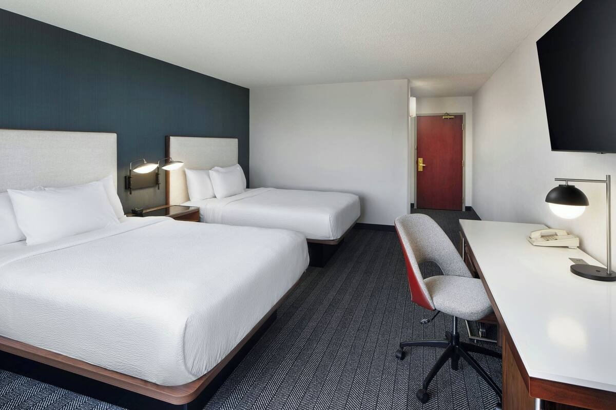 Quarto de hotel com duas camas, uma escrivaninha, uma cadeira e uma TV na parede.